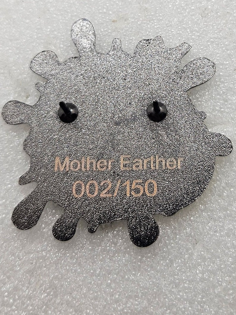 May include: Black enamel pin in the shape of a splatter with two black pin backs. The pin has the text "Mother Earth" and "002/150" in gold lettering.