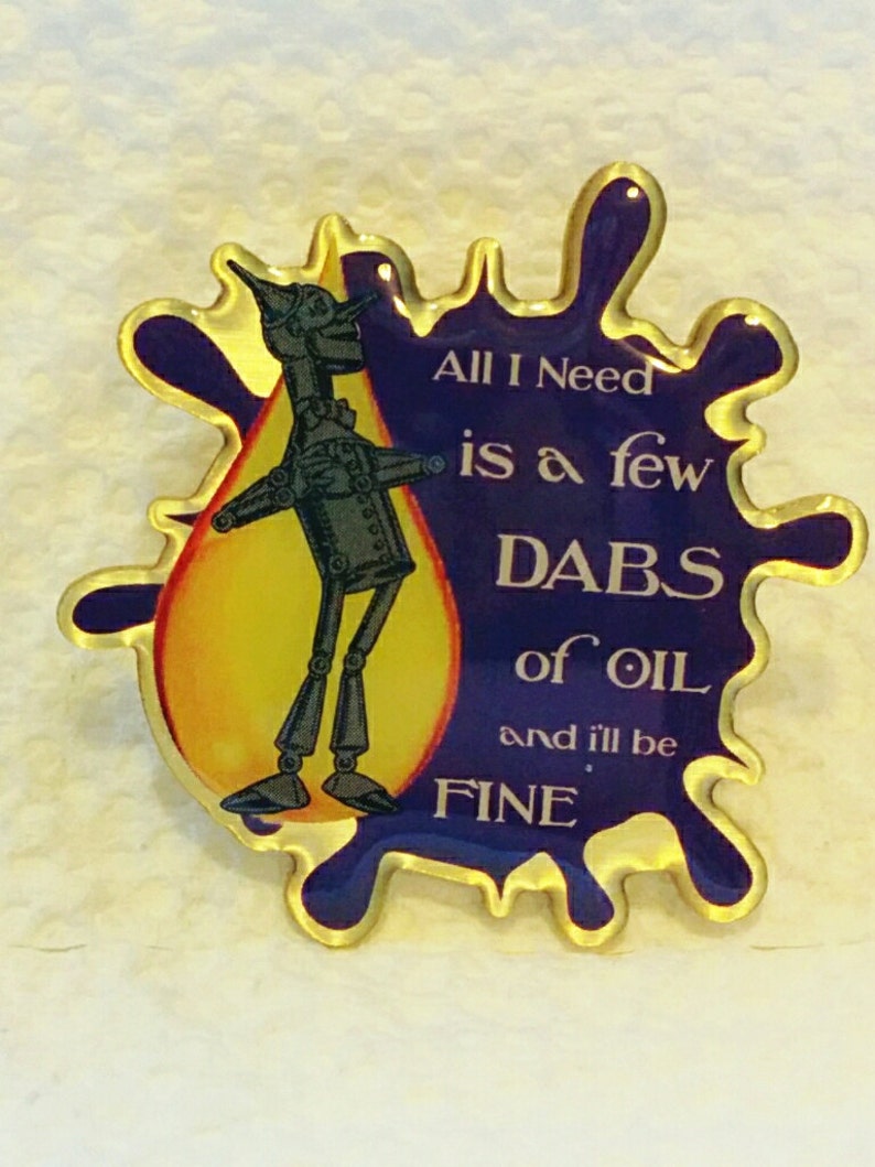 May include: A pin featuring the Tin Man from The Wizard of Oz. The pin has a gold border and a blue and yellow background. Text on the pin reads "All I Need is a few DABS of OIL and I'll be FINE."
