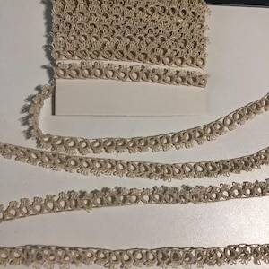 May include: A roll of beige lace trim with a repeating floral design. Several strands of the same lace are unrolled, showing the intricate details of the pattern. The lace is likely used for sewing, crafting, or adding decorative elements to garments or home decor.