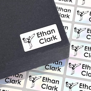 Puede incluir: Una hoja de etiquetas rectangulares blancas con una silueta negra de una persona dando una patada de karate. Las etiquetas tienen el nombre "Ethan Clark" impreso en texto negro.
