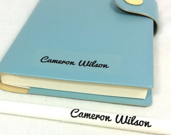 May include: A light blue notebook with a white label that says "Cameron Wilson" on it. A white pencil with the same name written on it is in front of the notebook.