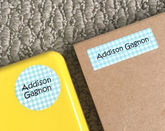Pode incluir: Duas etiquetas com padrão xadrez azul e branco com o nome "Addison Gagnon" impresso em preto. Uma etiqueta é redonda e está sobre uma superfície amarela. A outra etiqueta é retangular e está sobre uma superfície marrom.