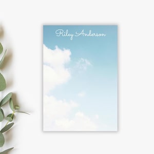May include: A notepad with a sky-themed design. The background features a gradient of light blue, with fluffy white clouds. The name "Riley Anderson" is written in elegant cursive at the top. The notepad is rectangular and sits on a white surface.