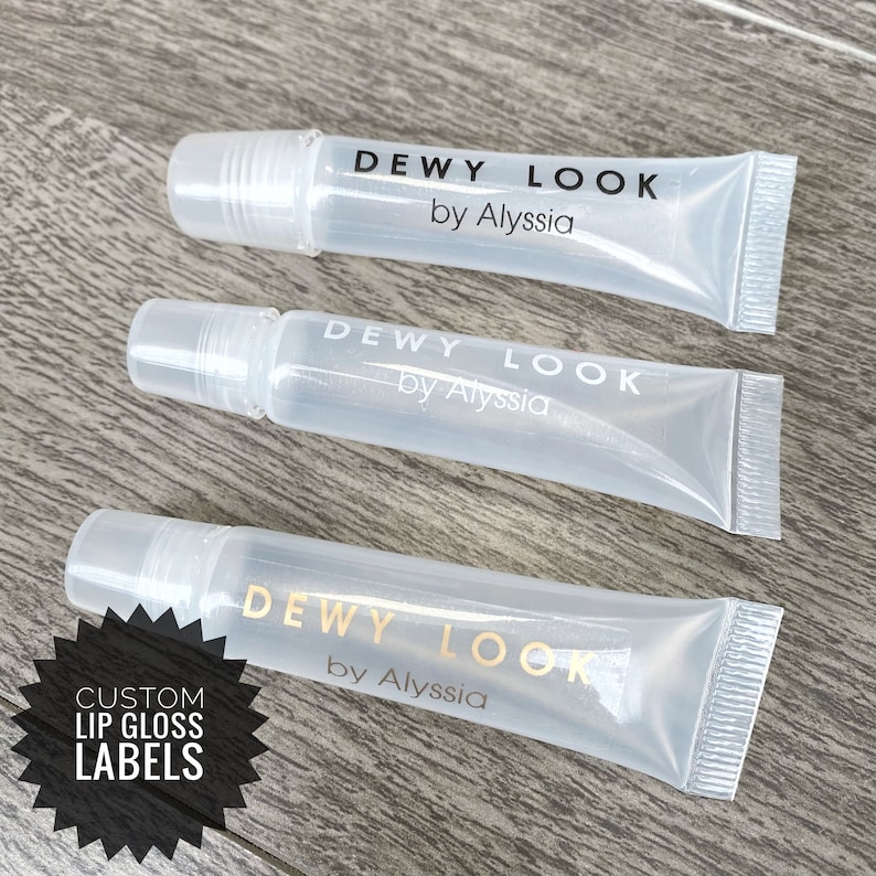 Puede incluir: Tres tubos de pl&aacute;stico transparente con etiquetas en blanco y negro que dicen "DEWY LOOK by Alyssia". Una pegatina dorada en forma de estrella con el texto "CUSTOM LIP GLOSS LABELS" est&aacute; en la esquina inferior izquierda de la imagen.