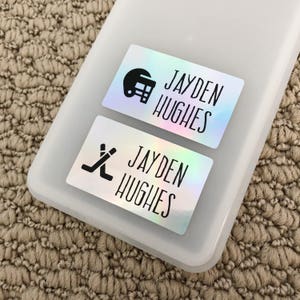 May include: Two iridescent labels with black text and icons. The top label reads "Jayden Hughes" and features a football helmet icon. The bottom label reads "Jayden Hughes" and features a hockey stick icon.