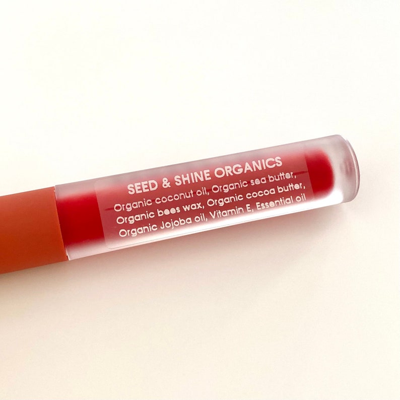 May include: A clear tube with a red liquid inside. The tube has a white label with the text "SEED & SHINE ORGANICS" and a list of ingredients: Organic coconut oil, Organic sea butter, Organic bees wax, Organic cocoa butter, Organic Jojoba oil, Vitamin E, Essential oil.