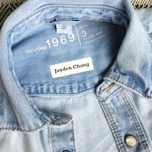 Peut inclure: Une chemise en jean bleu clair avec un col et des boutons-pression en métal. L'étiquette intérieure du col indique babyGap 1969 3 YEARS ANS. Une étiquette adhésive rectangulaire blanche est appliquée sur le tissu avec le nom Jayden Chang.