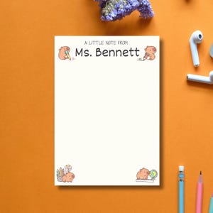 May include: A white notepad with the text "A Little Note From Ms. Bennett" and illustrations of cartoon pigs with pencils and a globe. The notepad is surrounded by pencils, earbuds, and flowers on an orange surface.