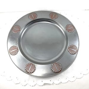 May include: A silver-colored plate with a raised rim, adorned with eight bronze-colored seashell designs. The plate has a smooth, reflective surface and is suitable for serving food. The design evokes a coastal or nautical theme.