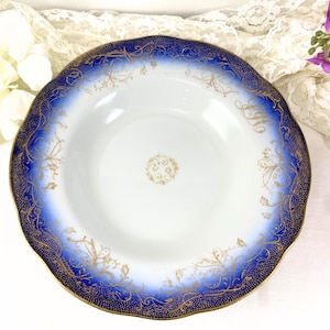 May include: A white ceramic bowl with a scalloped edge, featuring a deep blue border with gold filigree and a gold rim. The bowl's interior has a central gold floral design. A vintage decorative piece.