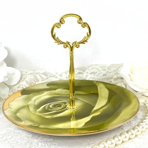 May include: A two-tiered serving tray with a gold handle and a floral design. The bottom tier features a large green rose, and the edges are trimmed in gold. The handle is ornate and gold-colored.