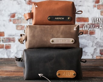 May include: Three leather toiletry bags stacked on top of each other. The top bag is brown and says "ANDREW" on it. The middle bag is beige and says "WILSON" on it. The bottom bag is black and says "AJH" on it. The bags are labeled with their sizes: "COMPACT (6 3/4 x 3 1/2 x 2 3/4)", "STANDARD (8 1/2 x 4 1/2 x 3 1/2)", and "LARGE (9 1/2 x 6 1/2 x 4 1/2)".