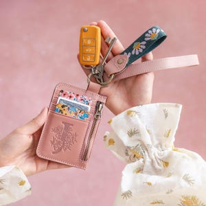 May include: A pink leather cardholder with a floral "J" design, a yellow car key, and a key fob with a daisy pattern. The cardholder has a zipper and holds several cards. A pink leather strap is attached to the key fob.