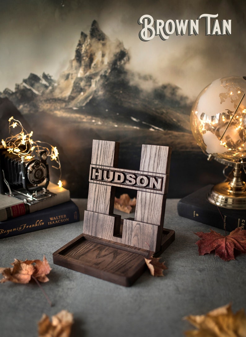 Pode incluir: Um suporte de telefone de madeira com um recorte para um telefone e uma pequena bandeja na parte inferior. O suporte &eacute; gravado com o nome "HUDSON" e tem um acabamento marrom escuro. O suporte est&aacute; sobre uma superf&iacute;cie cinza com algumas folhas de outono.