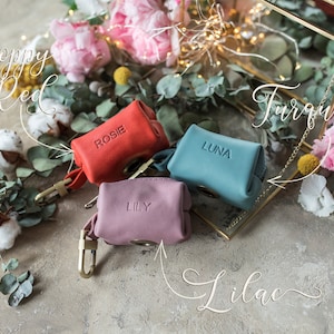 May include: Three small leather pouches with personalized names: Rosie, Luna, and Lily. The pouches are in red, turquoise, and lilac colors. The pouches are on a light brown surface with flowers and greenery.