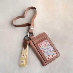 May include: A brown leather cardholder with a floral design and the text "Let your dreams blossom." It has a matching heart-shaped strap and a yellow daisy keychain. The cardholder is designed to hold cards and cash.