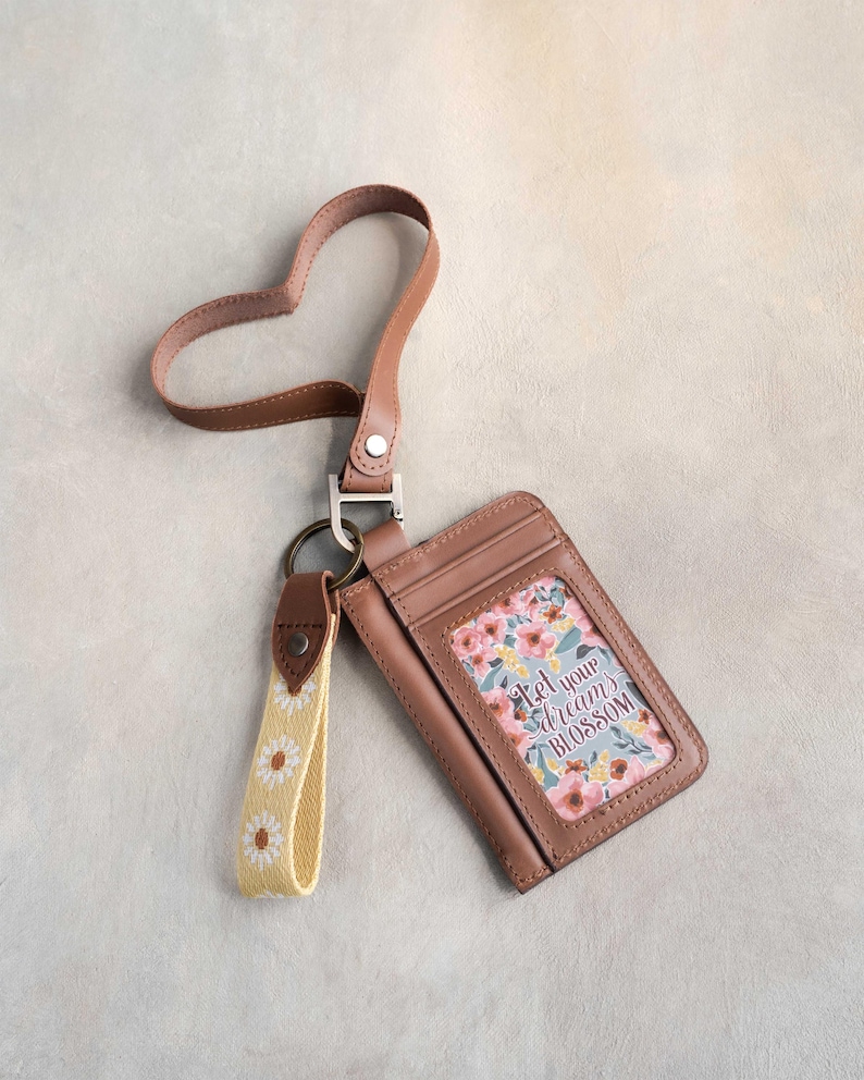 May include: A brown leather cardholder with a floral design and the text "Let your dreams blossom." It has a matching heart-shaped strap and a yellow daisy keychain. The cardholder is designed to hold cards and cash.