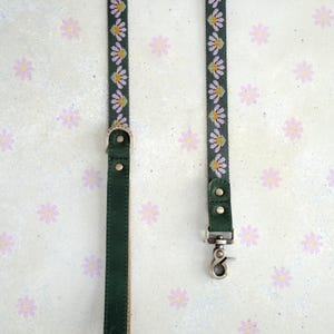 May include: Two dark green dog leashes with floral patterns. One leash has a silver clasp, and the other has a silver buckle. The floral pattern features white and yellow flowers on a dark green background. The background is a light color with small purple flowers.