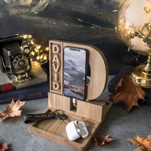 Pode incluir: Suporte de telefone de madeira com o nome "DAVID" gravado nele. O suporte tem um espa&ccedil;o para um telefone, um cabo de carregamento e uma bandeja pequena para chaves ou outros itens pequenos. O suporte &eacute; feito de madeira natural e tem um acabamento marrom claro. O suporte est&aacute; sentado em uma superf&iacute;cie cinza com algumas folhas de outono espalhadas ao redor. O texto "LOVE YOU DAD" est&aacute; em um chaveiro de couro marrom.