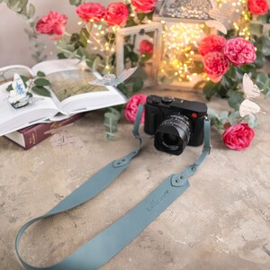 May include: A light blue leather camera strap with the word "Kalifornia" embossed on it. The strap is attached to a black camera with a lens. The camera is sitting on a light brown surface with pink flowers and a lantern in the background.