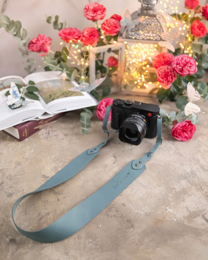 May include: A light blue leather camera strap with the word "Kalifornia" embossed on it. The strap is attached to a black camera with a lens. The camera is sitting on a light brown surface with pink flowers and a lantern in the background.