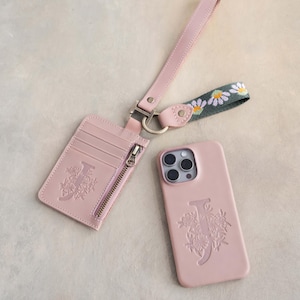 May include: A blush pink phone case, cardholder, and wrist strap set. The phone case and cardholder feature an embossed floral design with the letter "J". The wrist strap has a daisy chain pattern.