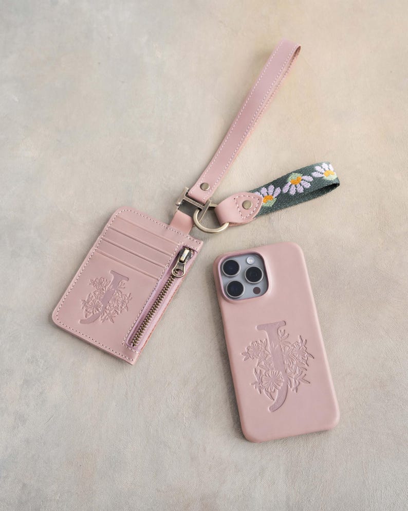 May include: A blush pink phone case, cardholder, and wrist strap set. The phone case and cardholder feature an embossed floral design with the letter "J". The wrist strap has a daisy chain pattern.