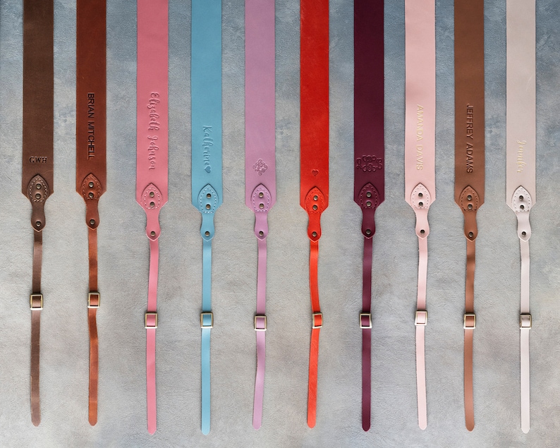 May include: Ten leather camera straps with personalized names in gold lettering. The straps are in various colors, including brown, pink, blue, red, and purple.