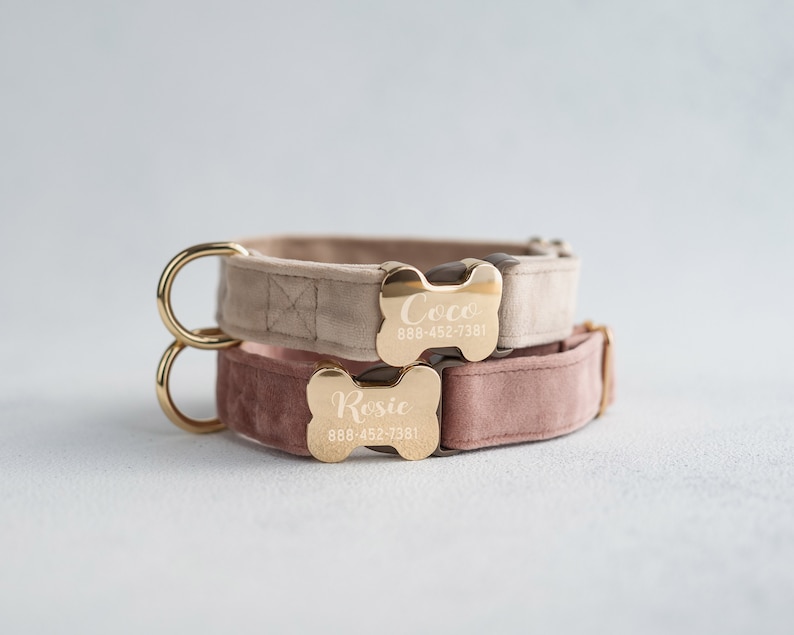 May include: Two dog collars with gold bone-shaped name tags. The top collar is beige velvet with the name "Coco" and phone number "888-452-7381" engraved on the tag. The bottom collar is pink velvet with the name "Rosie" and phone number "888-452-7381" engraved on the tag.
