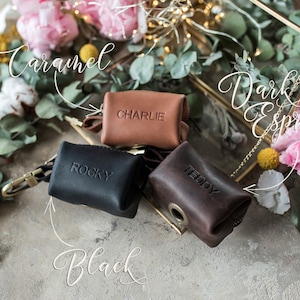 May include: Three small leather pouches with zipper closures and metal rings. The pouches are labeled "Charlie", "Rocky", and "Teddy". The pouches are displayed on a light brown surface with flowers and greenery.