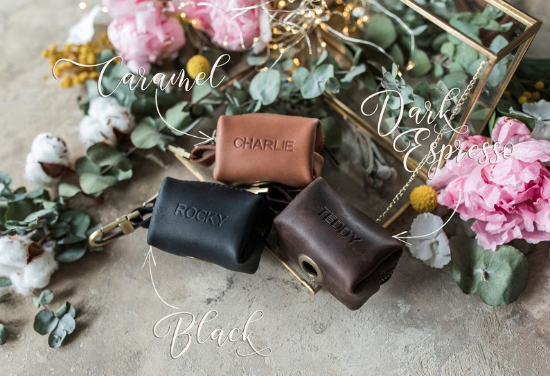 May include: Three small leather pouches with zipper closures and metal rings. The pouches are labeled "Charlie", "Rocky", and "Teddy". The pouches are displayed on a light brown surface with flowers and greenery.