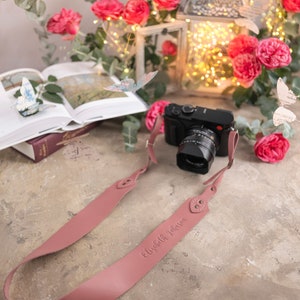 May include: A pink leather camera strap with the name "Elizabeth Johnson" embossed on it. The strap is lying on a light brown surface with a camera and a book in the background.