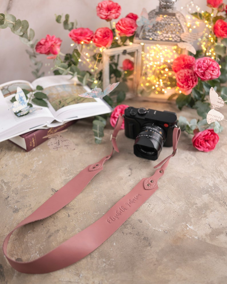 May include: A pink leather camera strap with the name "Elizabeth Johnson" embossed on it. The strap is lying on a light brown surface with a camera and a book in the background.