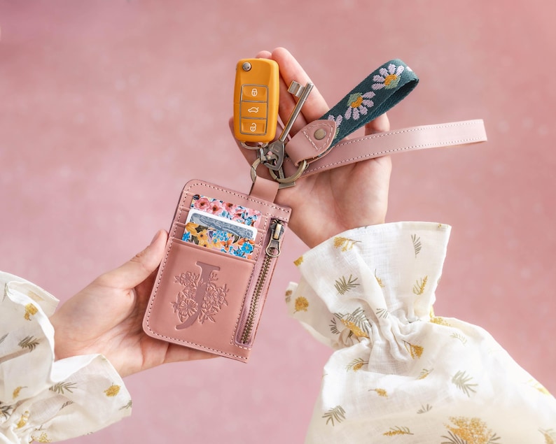 May include: A pink leather cardholder with a floral "J" design, a yellow car key, and a key fob with a daisy pattern. The cardholder has a zipper and holds several cards. A pink leather strap is attached to the key fob.