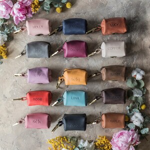 May include: Assortment of 14 small leather pouches with zipper closures and personalized names. The pouches are in various colors, including pink, blue, red, brown, beige, and black. The pouches are arranged in three rows on a textured gray surface.
