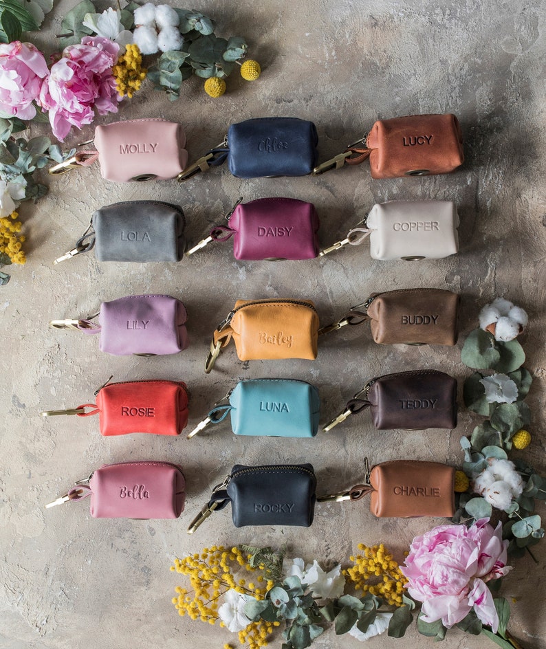 May include: Assortment of 14 small leather pouches with zipper closures and personalized names. The pouches are in various colors, including pink, blue, red, brown, beige, and black. The pouches are arranged in three rows on a textured gray surface.