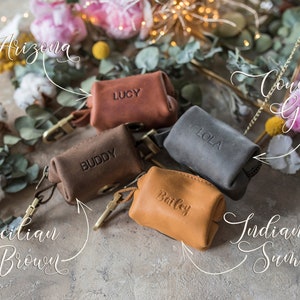 May include: Five personalized leather dog poop bag holders in different colors: Arizona, Sicilian Brown, Country Gray, Indian Summer, and Lucy. Each holder has a metal clip and a name engraved on the front.