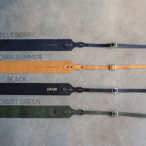 May include: Four leather camera straps in different colors: blueberry, indian summer, black, and forest green. Each strap has a brass buckle and a personalized name tag.