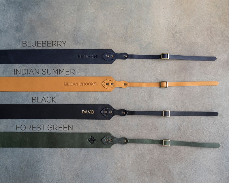 May include: Four leather camera straps in different colors: blueberry, indian summer, black, and forest green. Each strap has a brass buckle and a personalized name tag.