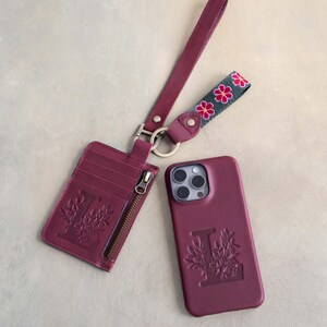 May include: A burgundy leather phone case, cardholder, and wrist strap set. The phone case and cardholder feature an embossed floral design with the letter "L". The wrist strap has a floral pattern in pink and green.