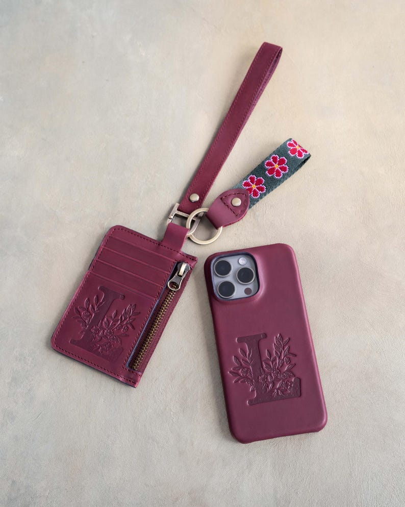 May include: A burgundy leather phone case, cardholder, and wrist strap set. The phone case and cardholder feature an embossed floral design with the letter "L". The wrist strap has a floral pattern in pink and green.