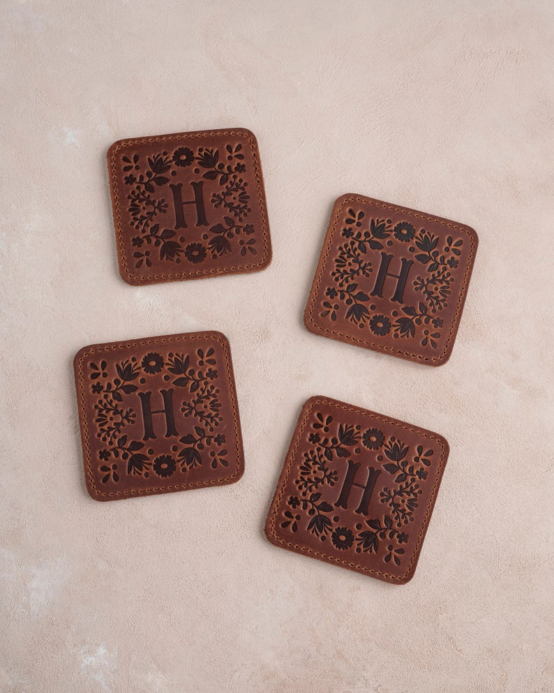 以下が含まれることがあります： 花柄のデザインと中央に「H」の文字が刻まれた、茶色の革製のコースターが4つあります。