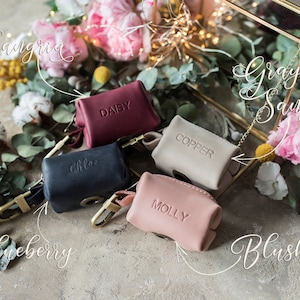 May include: Six small leather pouches in various colors, including navy blue, burgundy, beige, and pink. Each pouch has a gold metal clasp and a small metal tag with a name on it, such as "Daisy", "Chloe", "Copper", and "Molly". The pouches are arranged on a light brown surface with flowers and greenery in the background.