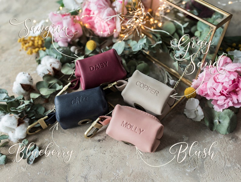 May include: Six small leather pouches in various colors, including navy blue, burgundy, beige, and pink. Each pouch has a gold metal clasp and a small metal tag with a name on it, such as "Daisy", "Chloe", "Copper", and "Molly". The pouches are arranged on a light brown surface with flowers and greenery in the background.