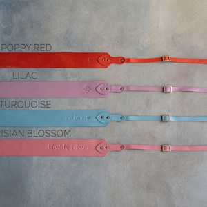 May include: Four leather camera straps in different colors: poppy red, lilac, turquoise, and Parisian blossom. Each strap has a gold buckle and a personalized name.
