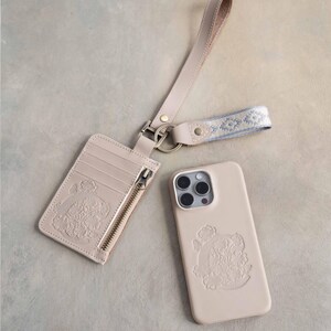 May include: A light gray cardholder, phone case, and wrist strap set. The cardholder has a zipper and slots, and the phone case has a camera cutout. All items feature an embossed floral design. The text "GRAY SAND" is at the top.