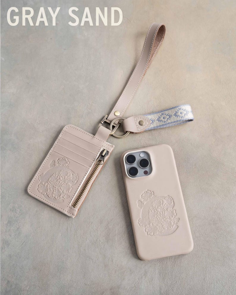 May include: A light gray cardholder, phone case, and wrist strap set. The cardholder has a zipper and slots, and the phone case has a camera cutout. All items feature an embossed floral design. The text "GRAY SAND" is at the top.