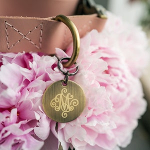 May include: A pink leather dog collar with a brass tag engraved with a monogram. The collar is resting on a bouquet of pink peonies.