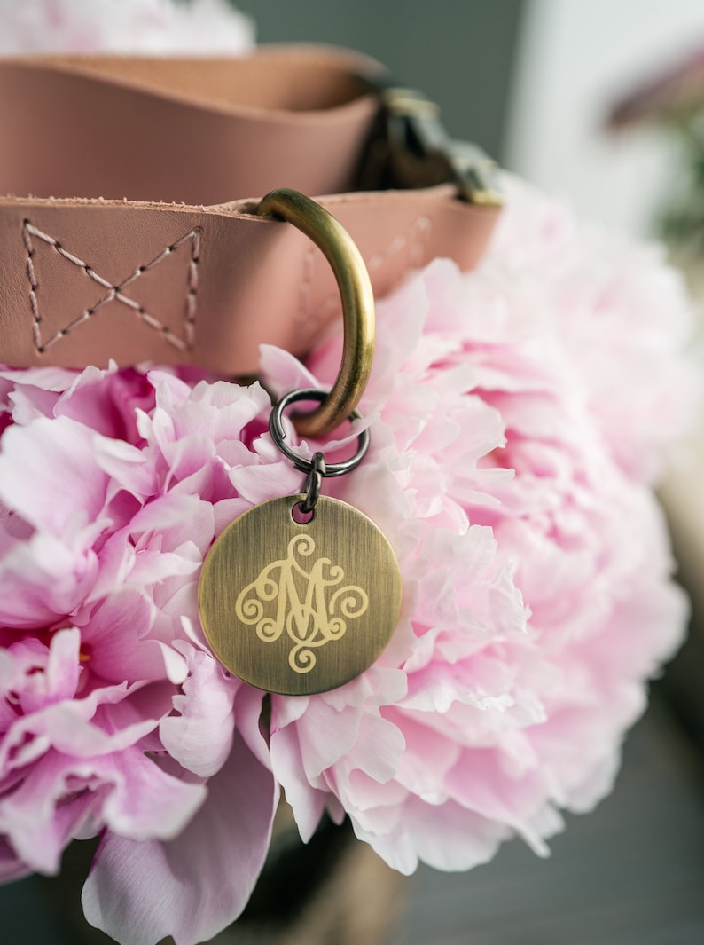 May include: A pink leather dog collar with a brass tag engraved with a monogram. The collar is resting on a bouquet of pink peonies.