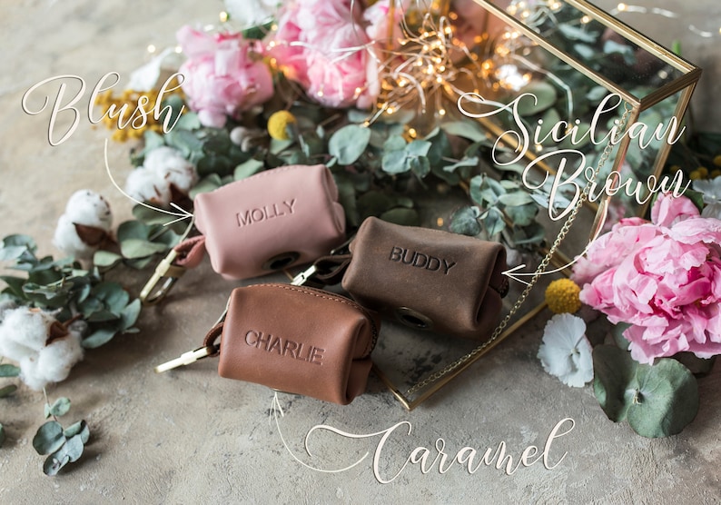 May include: Three small leather pouches with personalized names, "Molly", "Buddy", and "Charlie", in blush, caramel, and Sicilian brown colors. The pouches are surrounded by flowers and greenery.